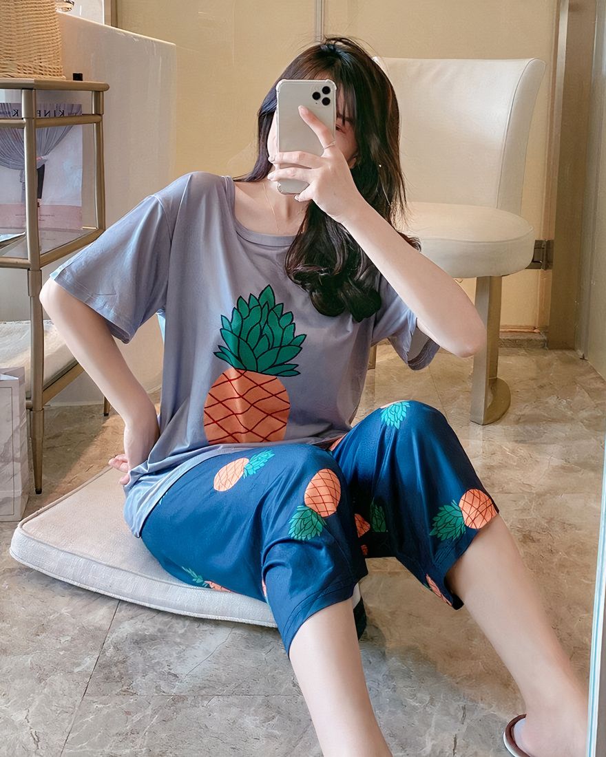 Two-piece pyjamas with pineapple motif worn by a woman sitting on a pillow in front of a mirror