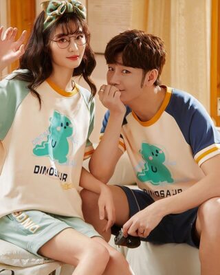 Two-piece cotton pajamas with t-shirt and shorts with dinosaur motif worn by a couple sitting on a bed in a house