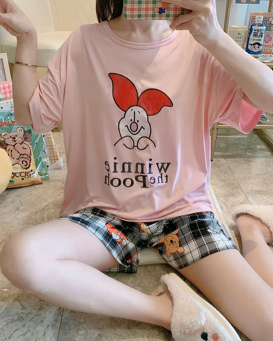 Pajama set with a pink t-shirt and shorts with a piglet pattern, worn by a woman sitting on a carpet in a house