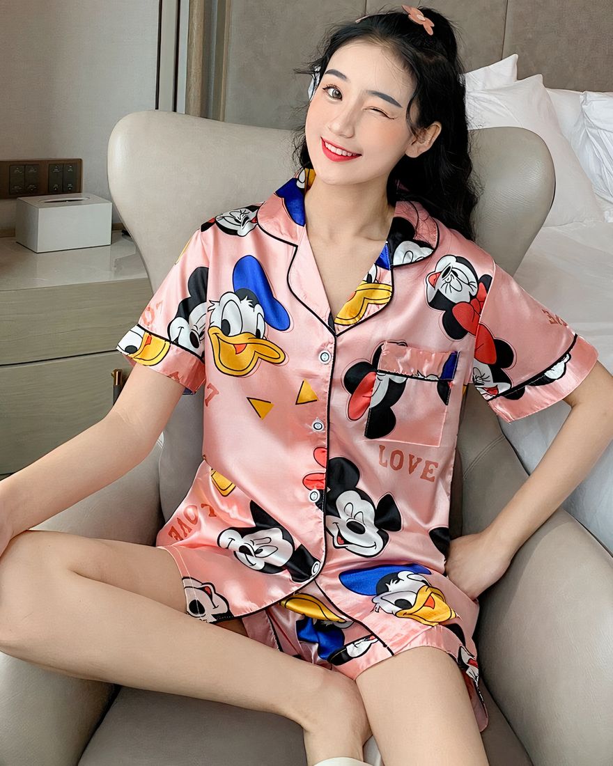 Two-piece pyjama set in pink satin with Mickey, Minnie and Donald motifs worn by a woman sitting on a chair in a house