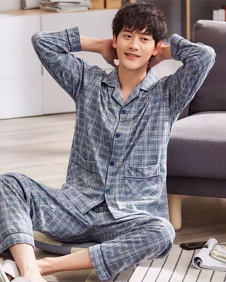 Men's pajamas in grey cotton stripes worn by a man sitting on a carpet in front of a sofa in a house