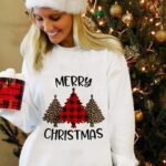 Women's long sleeve sweater with Christmas tree design and "Merry Christmas" worn by a woman in front of a Christmas tree in a house