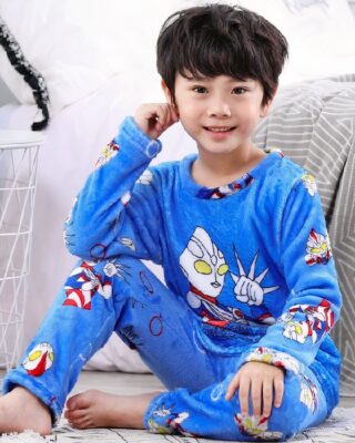 Blue boy's superhero flannel pajamas worn by a little boy sitting on a carpet in front of a bed in a house