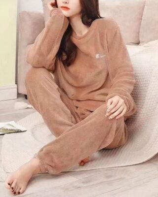 young woman wears a pyjama in beige pilou pilou, she is sitting on a plaid near a sofa, on the floor, the chin leaned in one of her hands